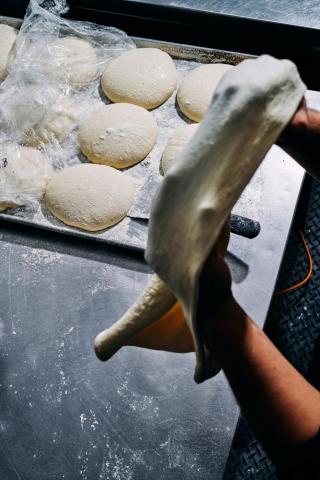 This image shows a person stretching pizza dough above a stainless steel table