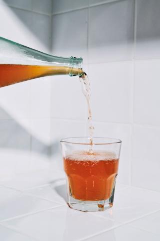 This image shows a clear glass bottle being tilted to pour a golden, fizzy liquid into a glass on a white tiled surface