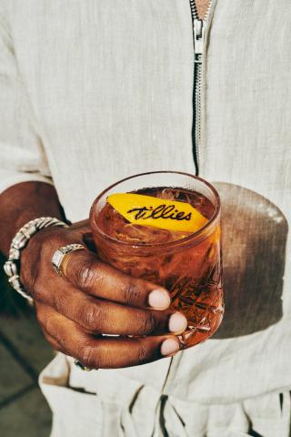 The image shows a close-up of a person wearing a white, zippered garment, holding an ornate glass of a chilled beverage, possibly a cocktail