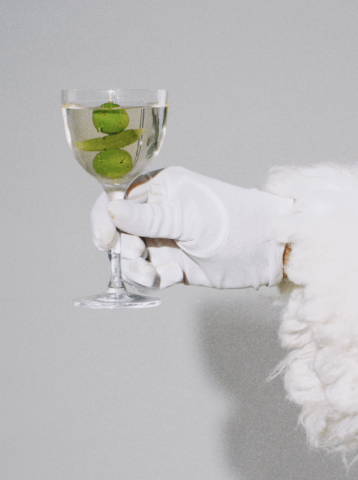 The image shows a hand wearing a white glove, holding a glass filled with clear liquid and three whole limes