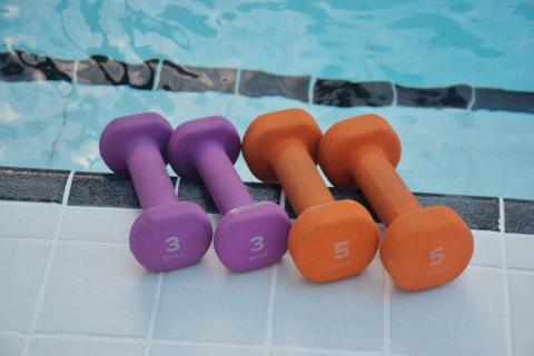 This image shows a set of colorful dumbbells, two pairs in purple and one pair in orange, resting on a white tiled surface by the edge of a pool