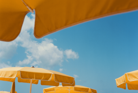 The image features several bright yellow beach umbrellas open against a vibrant blue sky with fluffy white clouds