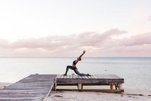 This image features a person performing a yoga pose on a wooden pier by the beach during what appears to be early morning or late afternoon