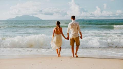 Couple walking on beach, man in shirt and shorts, woman in dress, ocean view.
