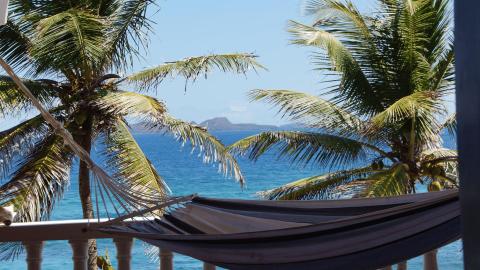 Serene beach view from a balcony with a hammock, palm trees, and distant island.