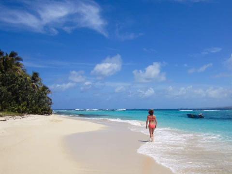 Person strolling on sandy beach with clear blue water, palm trees, and boat in the background.