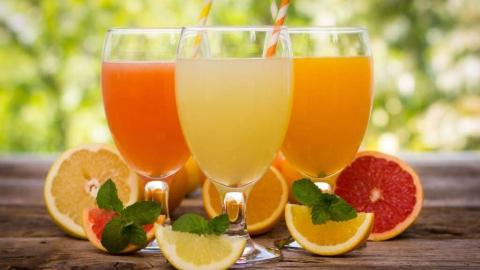 Three citrus juices in glasses with colorful straws, surrounded by sliced fruits and mint leaves.