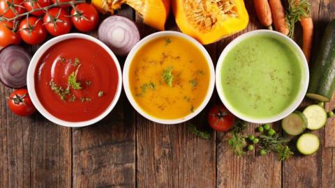 Three colorful bowls of soup with fresh vegetables on a wooden table.
