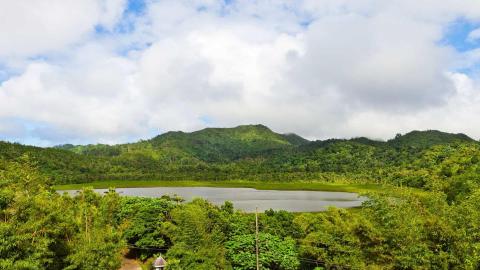 Tropical lake with lush forests, hills, and a thatched roof hut under partly cloudy skies.