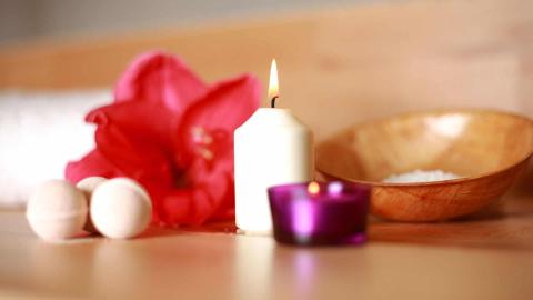 Spa setting with lit candles, red flower, salt bowl, and bath objects on wooden surface.