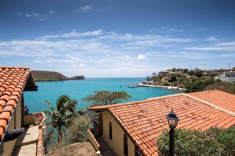 Coastal view with traditional roofs, palm trees, and anchored boats in a turquoise bay.