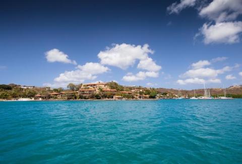 Coastal scene with turquoise sea, sailboats, green hills, and Mediterranean-style villas.