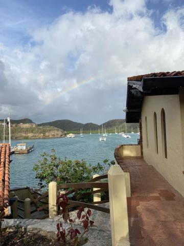Scenic waterfront view with boats, hills, a rainbow, and a terracotta roofed building.