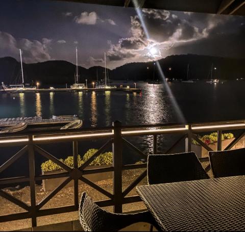 Serene marina night view with moonlight, boats, and mountains from a deck with chairs.