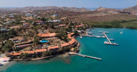 Aerial view of tropical harbor with moored boats, red-roofed buildings, and hilly backdrop