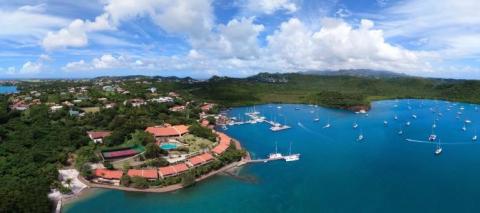 Aerial view of tropical island bay with boats, red-roofed buildings, and lush green hills