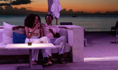 Two people toasting wine on a beach sofa at sunset, serene ocean view.