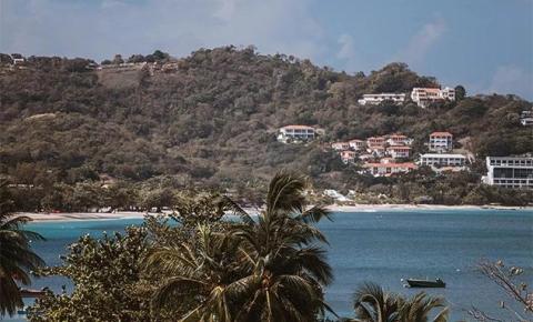 Coastal view with green hills, sandy beach, boats, and palm trees under a partly cloudy sky.