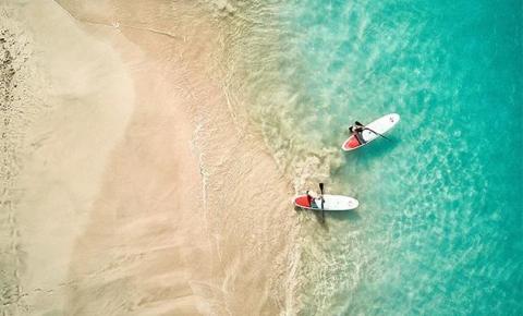 Aerial view of two paddleboarders on clear turquoise waters near sandy beach.