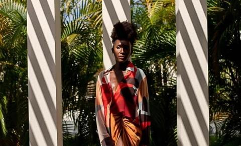 Woman in colorful shirt and mustard skirt stands between white pillars with green palms