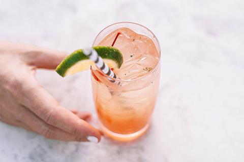 Hand holding a pink-orange drink with ice and a green citrus slice.