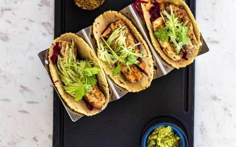 Three soft shell tacos with fried chicken, herbs, veggies, and green sauce on marble tray.