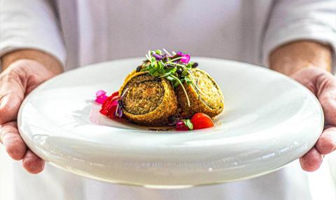 Chef presents gourmet stuffed roll with fresh greens and sauce on white plate.