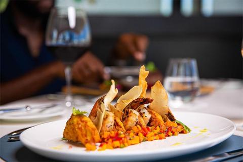 Grilled prawns on white plate with cracker, vegetables, and red wine in fine dining setting.