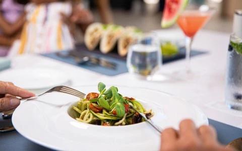 Person eating pasta with greens and tomato, tacos and cocktails in background