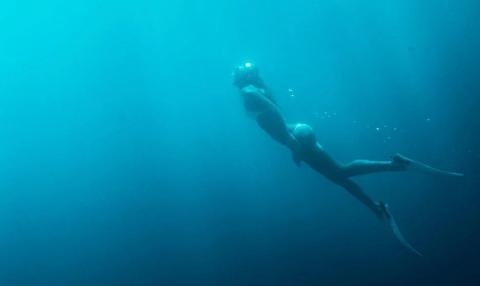 Underwater diver descending deep with fins and headlight in murky blue waters.