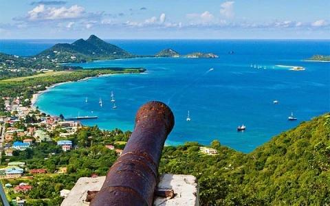 Old cannon overlooking a turquoise bay with sailboats and green mountains.