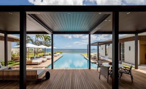 Luxurious poolside oasis extends to ocean under modern pergola with plush seating and palm trees.