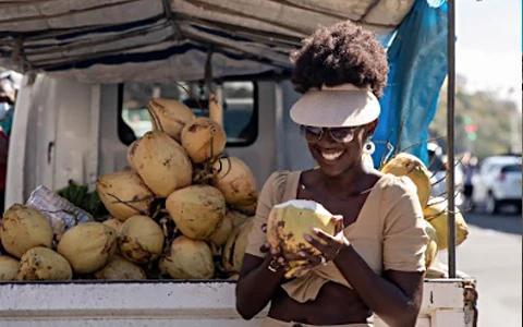 Woman drinking coconut at market stall, tropical street scene in background