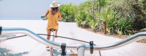 Woman in mustard dress walks with bike on tropical path, beach visible.