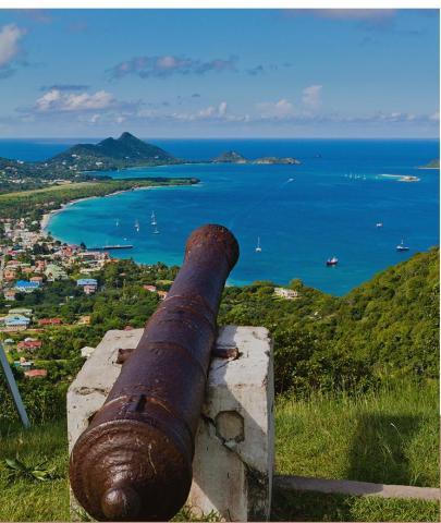 Old cannon overlooking coastal town with boats and green hills under clear sky