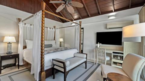 Spacious bedroom with canopy bed, seating area, desk, and TV in neutral tones.