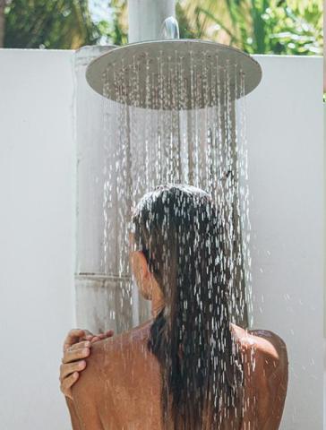 Female showering outdoors under sunlit showerhead with green leaves.