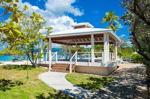 White gazebo by the sea surrounded by greenery and a paved path under a clear sky.