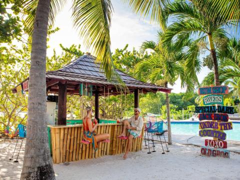 Tropical beach bar with palm trees, bamboo structure, and people relaxing.