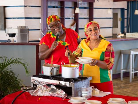 Two women laugh and serve food at a cultural cooking event indoors.