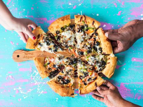 Colorful image of hands grabbing slices of a topped pizza on a peel