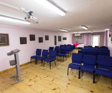 Interior meeting room with blue chairs, lectern, projector, and pale pink walls for gatherings.