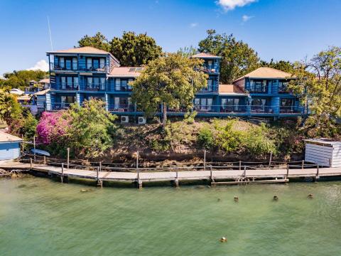 Elegant blue waterfront building with balconies, greenery, and a wooden dock