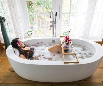 Woman in bubble bath with flowers in hair, bath tray with bouquets, in bright room