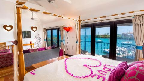 Romantic bedroom setup with ocean view, heart-shaped petals on bed, scenic balcony view.