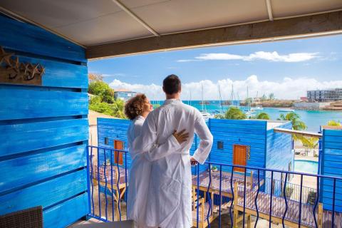 Couple in robes on balcony overlooking harbor with sailboats and construction.