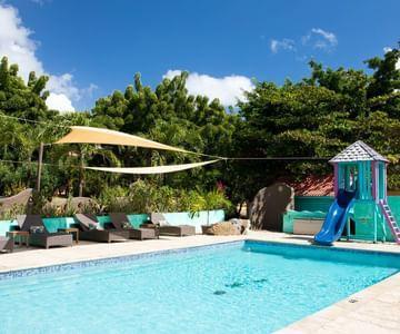 Outdoor pool with slide, sun loungers, and yellow canopy surrounded by greenery.