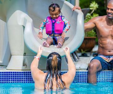 Child sliding down water slide into pool with two adults waiting to catch her.