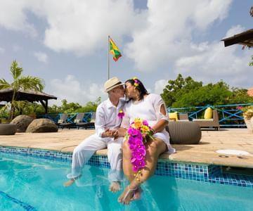 Older couple kissing poolside in resort, man in white suit, woman in floral dress with flowers.