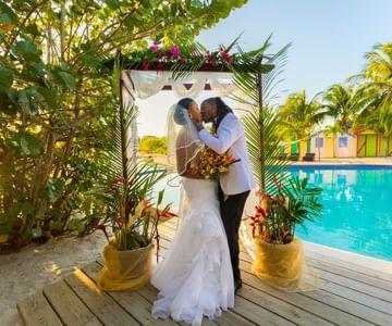 Couple embracing under floral arch by pool at tropical wedding.
